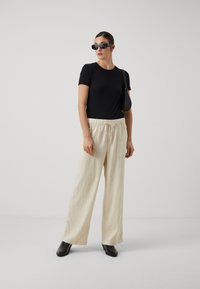 T-shirt nero aderente, pantaloni larghi crema con cintura elastica e tasche, e stivali neri con tacco spesso. Modella che indossa occhiali da sole.
