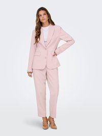 Sieviete ar gariem viļņainiem matiem, tērpusies gaiši rozā stilīgā uzvalkā, baltā topā un bēšos augstpapēžu kurpēs, stāvot ar vienu roku uz gurniem.