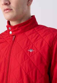 Rode gevoerde jas met hoge kraag, met drukknopen en een logo op de borst. De stof lijkt glad met een gestructureerde afwerking.