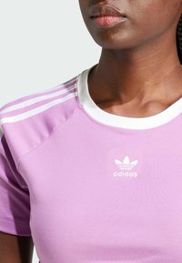 Lilac short-sleeve t-shirt made of smooth fabric, featuring a white crew neck and three white stripes on each shoulder with an adidas logo.