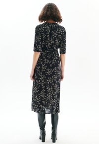 Femme aux cheveux bouclés portant une robe midi noire à fleurs et des bottes noires à talons jusqu'aux genoux, se tenant dos au spectateur.