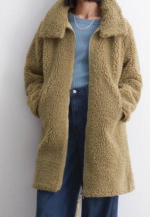 Cappotto in felpa khaki con una superficie texturizzata e soffice, dotato di collo, chiusura con zip e tasche laterali, indossato sopra una maglietta blu.