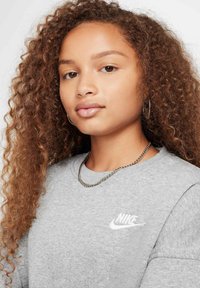 Szary sweter z długimi rękawami i okrągłym dekoltem. Posiada białe logo Nike na klatce piersiowej. Noszony z srebrnym naszyjnikiem.