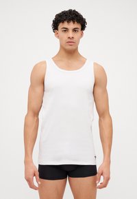 Λευκή αμάνικη μπλούζα, με ριγέ υφή, στρογγυλή λαιμόκοψη και φαρδιές τιράντες. Φοριέται με μαύρα κοντά σορτς, αναδεικνύοντας τα χέρια και τον κορμό.