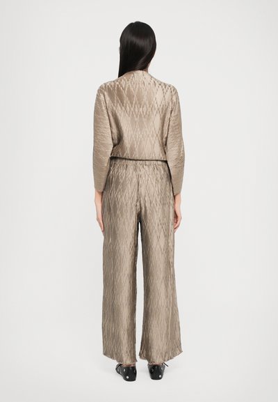 Beige, plissert jumpsuit med vid bendesign, belte i midjen. Tekstuert stoff med et subtilt mønster. Lange ermer og en komfortabel passform.
