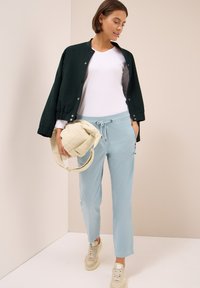 Veste bomber vert foncé, tee-shirt blanc à col en V, pantalon bleu clair à cordon, sac beige et baskets beiges avec semelles texturées.