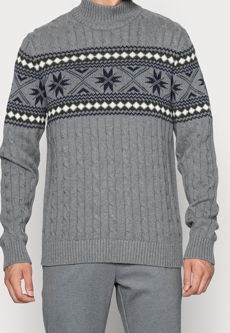 Homme portant un pull en maille torsadée gris avec une bande à motifs de formes géométriques et d'étoiles sur la poitrine et le haut des manches.