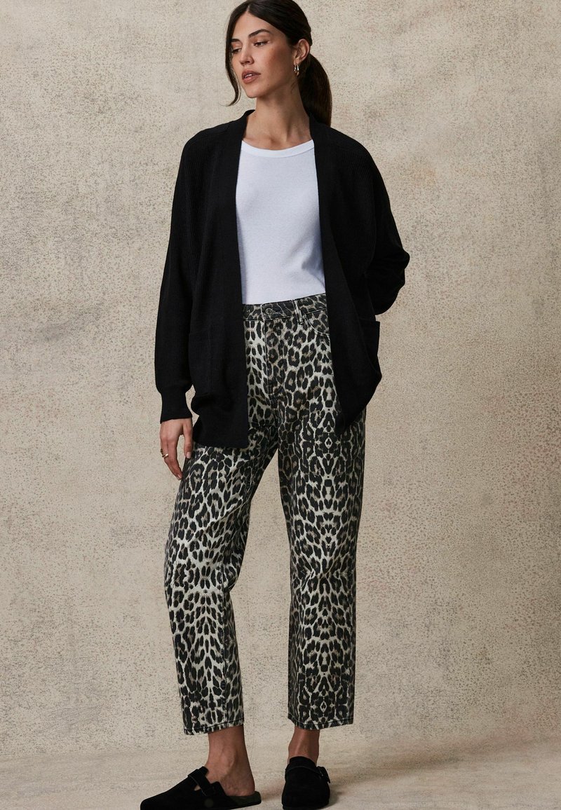 Schwarzer gerippter Cardigan und weißes enges T-Shirt kombiniert mit gemusterten grauen und schwarzen Leopardenprint-Hosen. Schwarze Slipper.