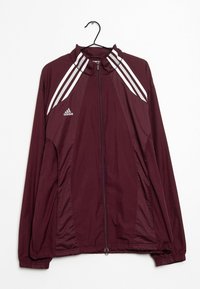 Bordeaux Adidas zip-up jack met witte strepen op de schouders, elastische mouwen en tailleband, hangend aan een zwarte hanger tegen een witte achtergrond.