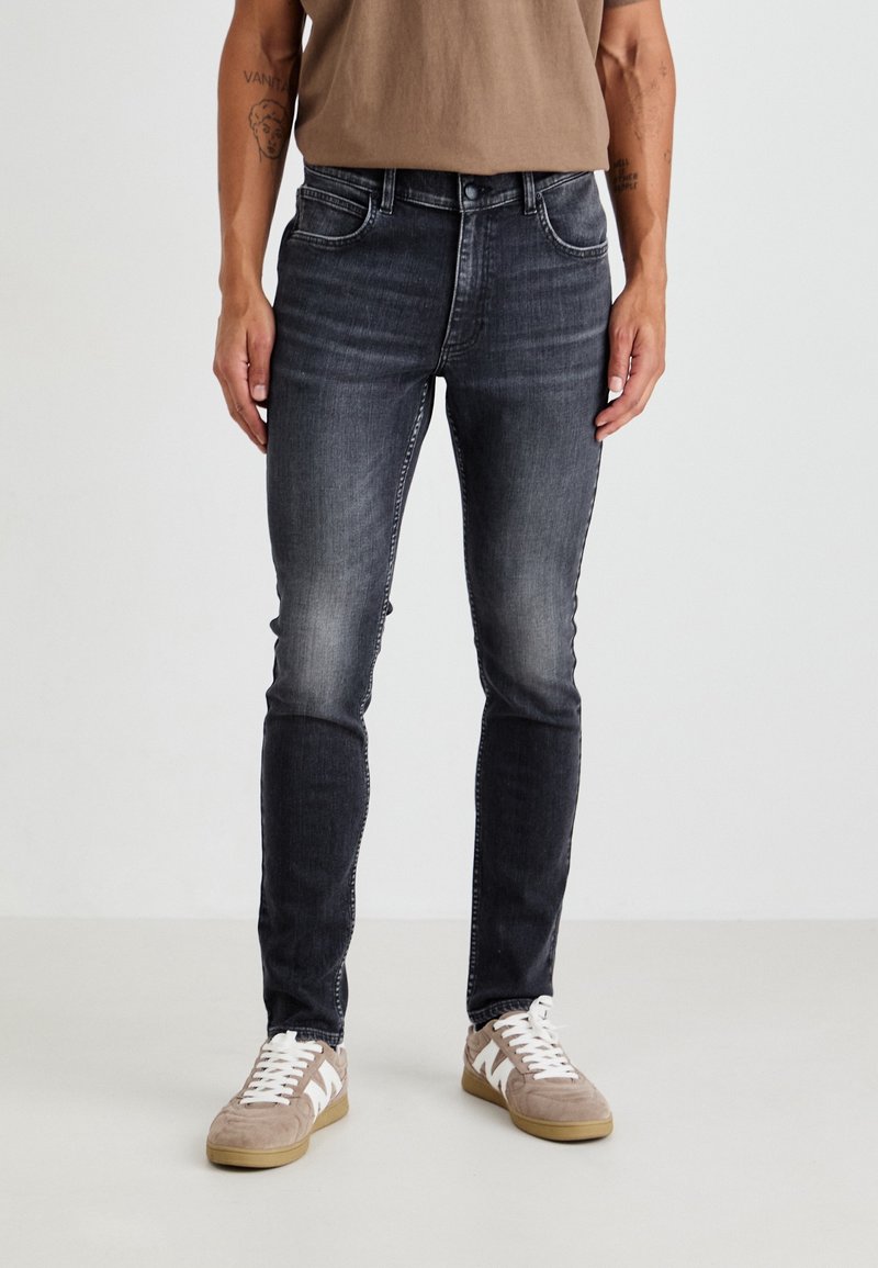 Homme portant un jean noir délavé slim fit et des baskets beige, debout devant un fond uni avec les mains relâchées le long du corps.