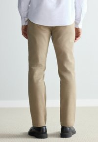 Beige cotton trousers with a slim fit, straight legs, and hemmed cuffs, paired with black textured shoes, viewed from the back.