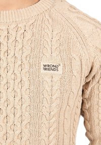 Beige stickad tröja med ett texturerat kabelmönster, ribbad halsringning och en liten "WRONG FRIENDS" etikett på vänster framsida.