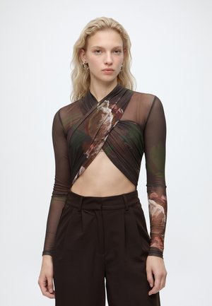 Jeune femme aux longs cheveux blonds porte un top court transparent à fleurs et un pantalon marron foncé taille haute sur un fond uni.