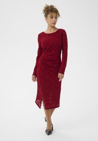 Abito rosso di pizzo con maniche lunghe, motivo testurizzato, spacco laterale per facilitare il movimento, abbinato a décolleté nere con punta a stiletto.