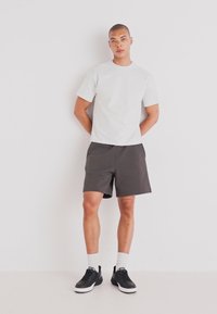 Grå t-skjorte og mørke shorts laget av stoff. Modellen har på seg svarte sneakers med hvite såler og hvite ankelsokker. Minimalistisk design.
