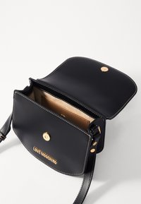 Black leather crossbody bag with a gold snap closure, contrasting white stitching, and embossed logo on the front. Interior is tan fabric.