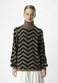 Maglione a collo alto con motivo a zigzag marrone e nero, con maniche larghe, texture morbida e vestibilità rilassata, indossato sopra una gonna bianca.
