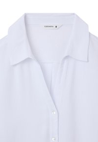 Camicia bianca con colletto, realizzata in tessuto morbido. Presenta una patta con piccoli bottoni rotondi e un'etichetta all'interno del colletto.