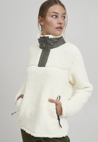 Crèmekleurige fleece pullover met een hoge kraag en een groene ritssluiting aan de voorkant. Inclusief zijzakken met trekkoorddetails. Zachte textuur.
