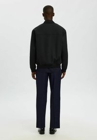 Giacca nera con zip, colletto e polsini a costine, indossata con pantaloni blu navy. Il tessuto appare liscio, senza motivi o accessori visibili.