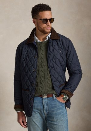 Homme portant une veste matelassée foncée par-dessus un pull vert à torsades, jean bleu clair, lunettes de soleil et montre, posant avec une main dans la poche.