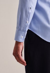 Jasnoniebieska koszula w kratę z białymi guzikami i haftowanym akcentem. Manżeta ma zaokrąglony brzeg. Gładka tekstura, dopasowany krój.