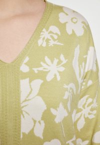 Helles grünes Strickpullover mit V-Ausschnitt und weißen Blumenmustern. Der strukturierte Rand entlang des Ausschnitts verleiht dem Design zusätzliche Details.