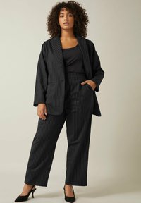 Zwarte pinstripe blazer en broek van een gladde stof. De blazer heeft een open voorkant en twee zakken; de broek heeft een rechte pijp en een hoge taille.