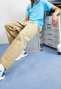 T-shirt en coton bleu clair, pantalon beige décontracté avec logo, baskets multicolores à semelle épaisse, le tout sur un sol bleu clair et une imprimante.