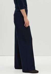 Pantalons larges bleu marine avec une texture lisse, dotés d'une taille mi-haute et de poches latérales. Le tissu tombe élégamment jusqu'au sol.