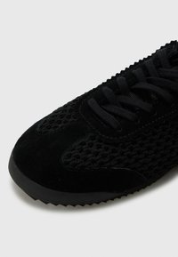 Scarpa sportiva nera caratterizzata da una combinazione di suede liscia e materiali a rete, con una superficie texturizzata, lacci piatti e una suola in gomma.