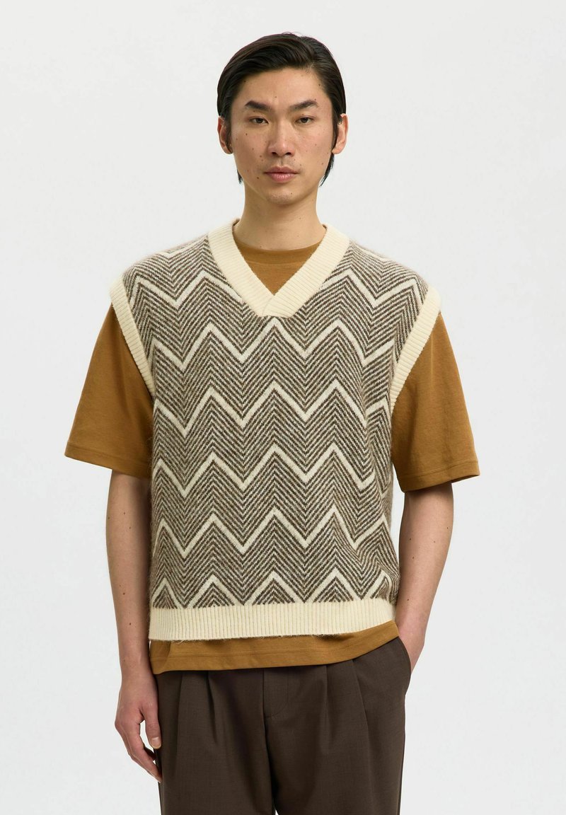 Gilet en mélange de laine, crème et marron à motif zigzag, avec un col en V profond, superposé sur un T-shirt marron, assorti à un pantalon marron.