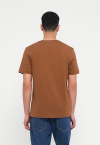 Καφέ κοντομάνικο μπλουζάκι με στρογγυλή λαιμόκοψη και λεία υφή, συνδυασμένο με μπλε τζιν παντελόνι, φαίνεται από πίσω.