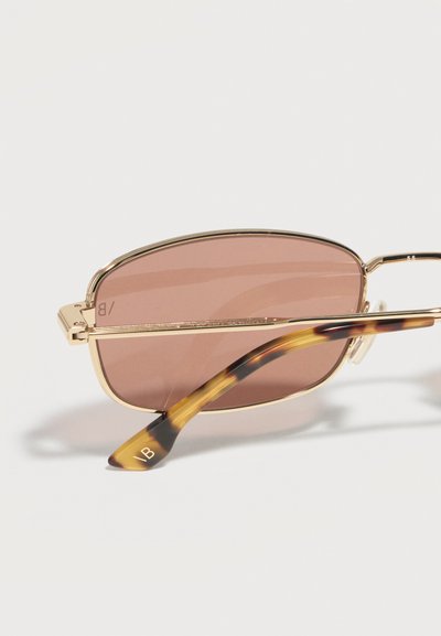 Γυαλιά ηλίου με χρυσή κορνίζα, ορθογώνια καφέ φακούς με σκίαση και πλαίσια με μοτίβο χελώνας, ξεκουράζονται σε λευκή επιφάνεια.