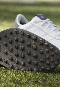 Scarpa sportiva bianca con accenti grigi; suola in gomma nera testurizzata con distinti stud chetoni per una migliore aderenza su superfici erbose.