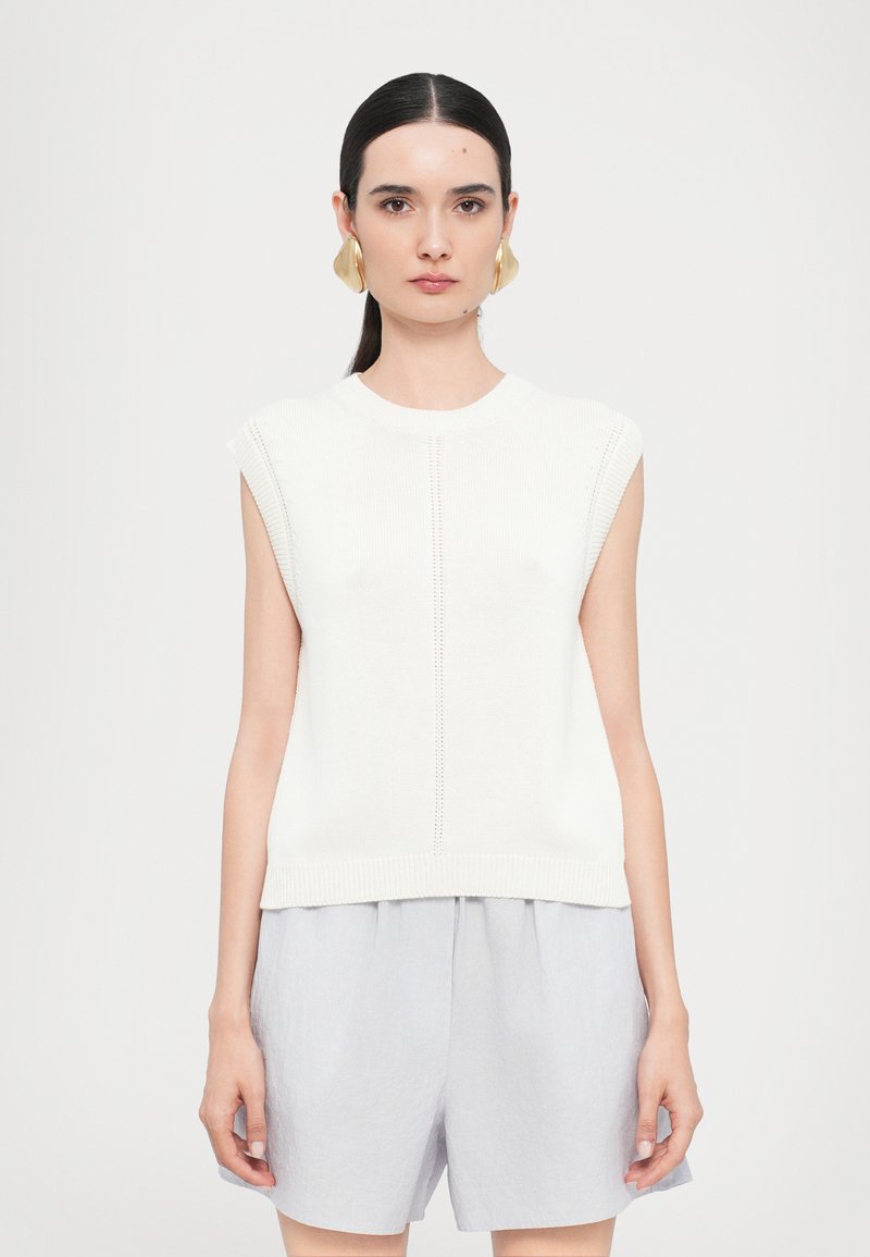 Top senza maniche in maglia bianca con scollatura tonda, caratterizzato da un dettaglio a linea centrale. Abbinato a pantaloni corti di colore grigio chiaro, in tessuto leggero e ampio.