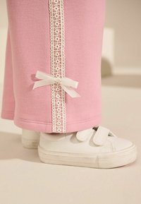 Rosa Jogginghosen mit weißer Spitzenkante und Schleifchen-Detail am Knöchel, kombiniert mit weißen Klettsneakern. Die weiche Stoffstruktur ist sichtbar.