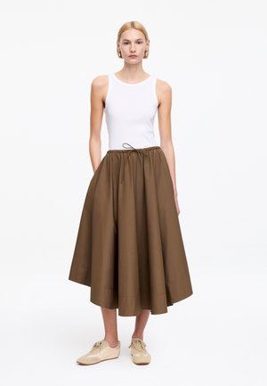 Blondynka w białym bezrękawniku, brązowej spódnicy o długości do połowy łydki, rozkloszowanej z wiązaniem w talii, oraz beżowych trampkach, stojąca na białym tle.