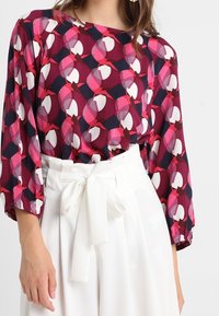 Blouse à manches longues présentant un motif géométrique dans des nuances de rose, bordeaux et marine sur un tissu léger. Associée à un pantalon blanc taille haute.