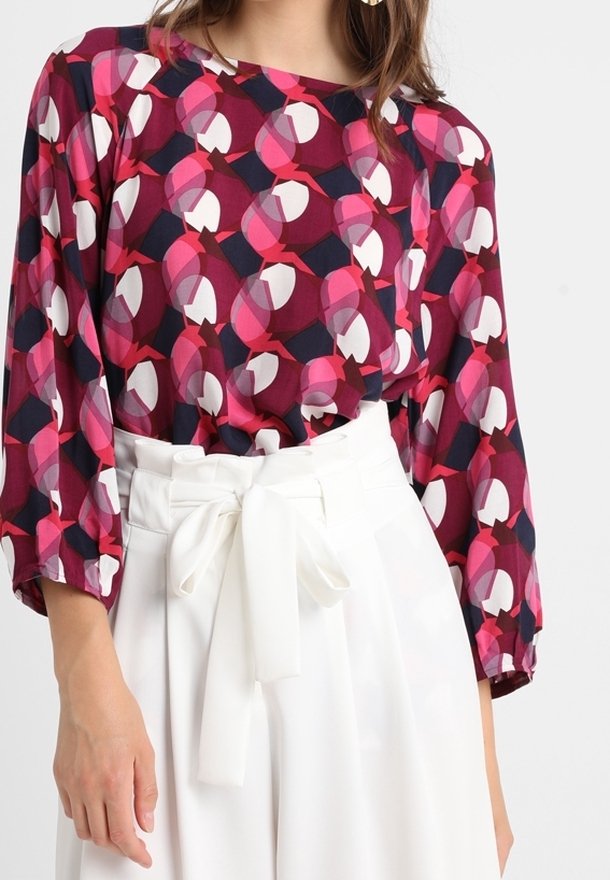 Blouse à manches longues présentant un motif géométrique dans des nuances de rose, bordeaux et marine sur un tissu léger. Associée à un pantalon blanc taille haute.