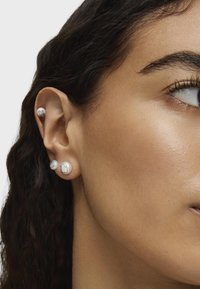Orecchini a perno in argento e perla con un design rotondo e liscio, caratterizzati da una superficie testurizzata. Sono visibili più fori di piercing nell'orecchio.