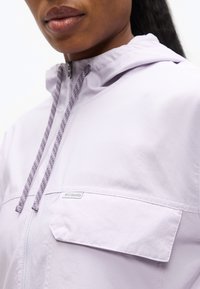 Κοντινό πλάνο ατόμου που φοράει ανοιχτόμωβ μπουφάν Columbia με φερμουάρ, κουκούλα και κορδόνια, εμφανίζοντας μερικώς τα χείλη και το κάτω μέρος του προσώπου.