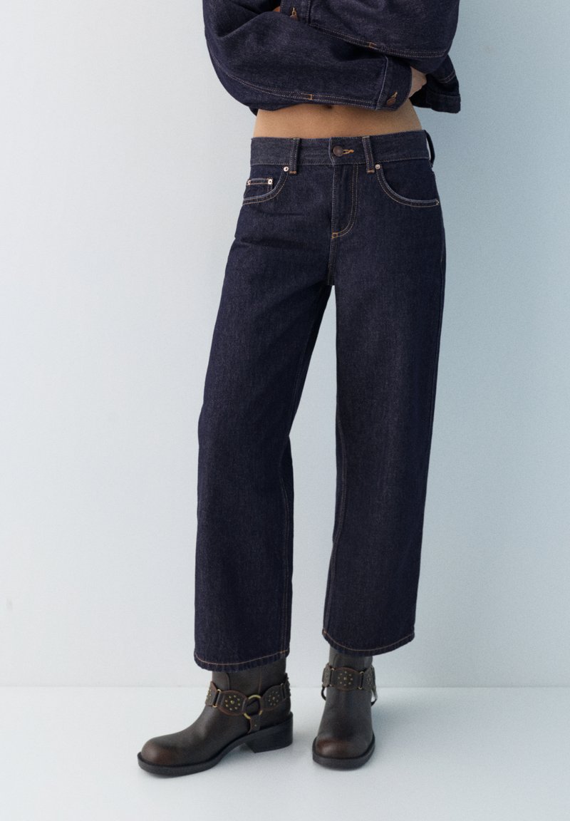 Persona che indossa jeans scuri a gamba larga, stivaletti neri con fibbie e una giacca scura corta, su uno sfondo chiaro e uniforme.