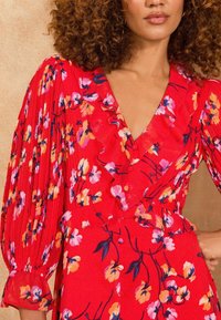 Rochie florală roșie cu decolteu în V și detalii cu volane. Prezintă mâneci lungi, plisate și o talie ajustată cu modele colorate de flori.