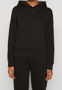 Felpa nera con cappuccio, in morbido cotone misto, vestibilità comoda, con toppe logo frontale, polsini e orlo a costine, texture liscia. Indossata con pantaloni abbinati.
