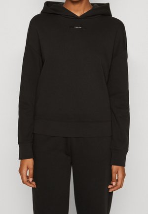 Sweatshirt à capuche noire, en mélange de coton doux, coupe décontractée, patch logo sur le devant, poignets et ourlet côtelés, texture lisse. Porté avec un pantalon assorti.