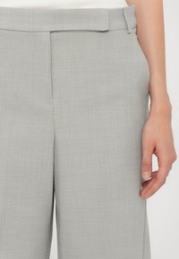 Gros plan d’un pantalon gris clair sur mesure avec une poche latérale et une main droite visible sur fond blanc.