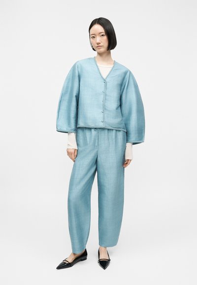 Γυναίκα που φοράει άνετο, ανοιχτό μπλε κοστούμι από λινό με φαρδιά μανίκια, συνδυασμένο με λευκή εσωτερική μπλούζα και μαύρα μυτερά φλατ παπούτσια.
