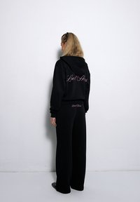 Schwarzer Kapuzenpullover und weite Hose mit Logo-Detail auf der Rückseite und der Seite. Weicher Stoff mit lässiger Passform und nahtloser Verarbeitung.