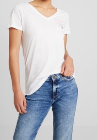 Camiseta de algodón blanca con escote en V y mangas cortas. Presenta "TOMMY JEANS" impreso en rosa. Combinada con jeans de mezclilla azul.